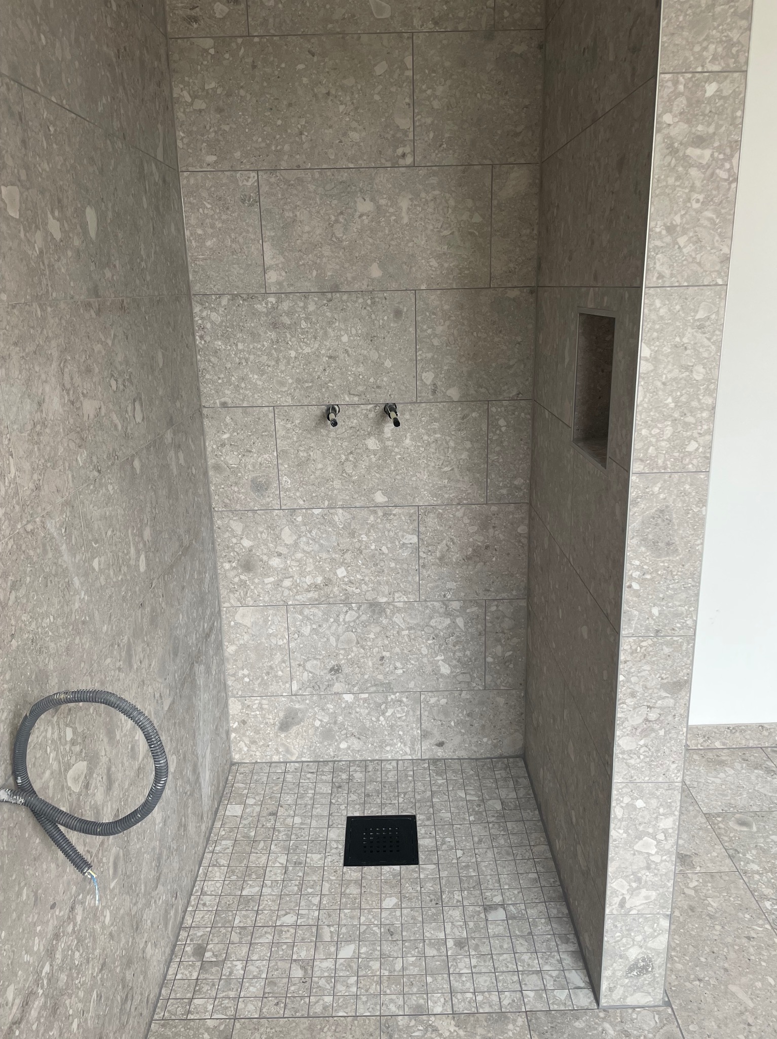 Wykończona kabina prysznicowa z szarymi płytkami imitującymi lastryko, widoczny odpływ liniowy, wnęka w ścianie i wystające rury.