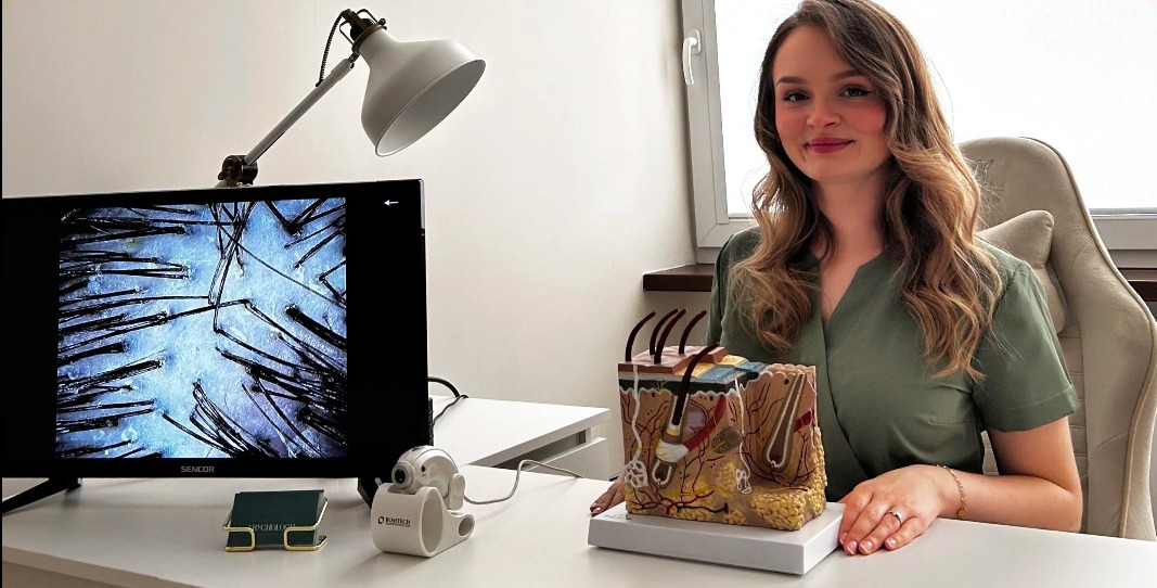 Trycholog prezentuje obraz skóry głowy na monitorze, obok model skóry i urządzenie diagnostyczne. Kobieta w zielonym uniformie uśmiecha się do kamery.