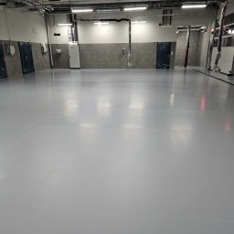 FLOORIGO - Jasnoszara, gładka posadzka w hali przemysłowej z widocznymi instalacjami pod sufitem i drzwiami w tle. Minimalistyczny design, czysta powierzchnia.