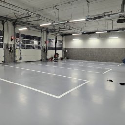 FLOORIGO - Szara, gładka podłoga w hali z wyznaczonymi białymi liniami stanowisk. Widoczne bramy garażowe, oświetlenie sufitowe i instalacje. Minimalistyczny, przemysłowy styl.