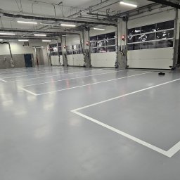 FLOORIGO - Jasnoszara posadzka przemysłowa z białymi liniami wyznaczającymi miejsca parkingowe w hali z bramami segmentowymi i instalacjami pod sufitem.