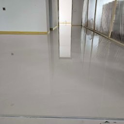 FLOORIGO - Błyszcząca, jasnoszara posadzka w nowym wnętrzu. Widoczne zabezpieczenia ścian folią i taśmą. Gładka powierzchnia odbija światło, tworząc efekt lustra.
