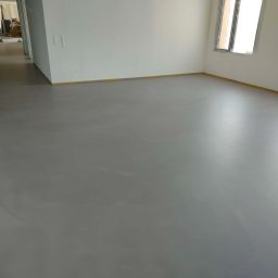 FLOORIGO - Szara, gładka podłoga w jasnym pomieszczeniu z oknami zabezpieczonymi folią. Żółta taśma malarska wzdłuż ścian. Widok na dalsze pomieszczenia w głębi.