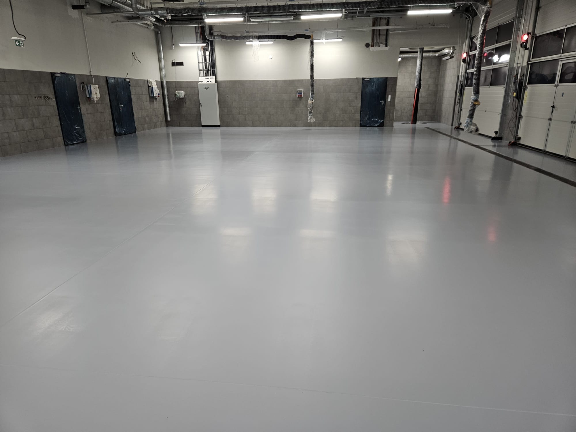 Jasnoszara, gładka posadzka w hali przemysłowej z widocznymi instalacjami pod sufitem i drzwiami w tle. Minimalistyczny design, czysta powierzchnia.