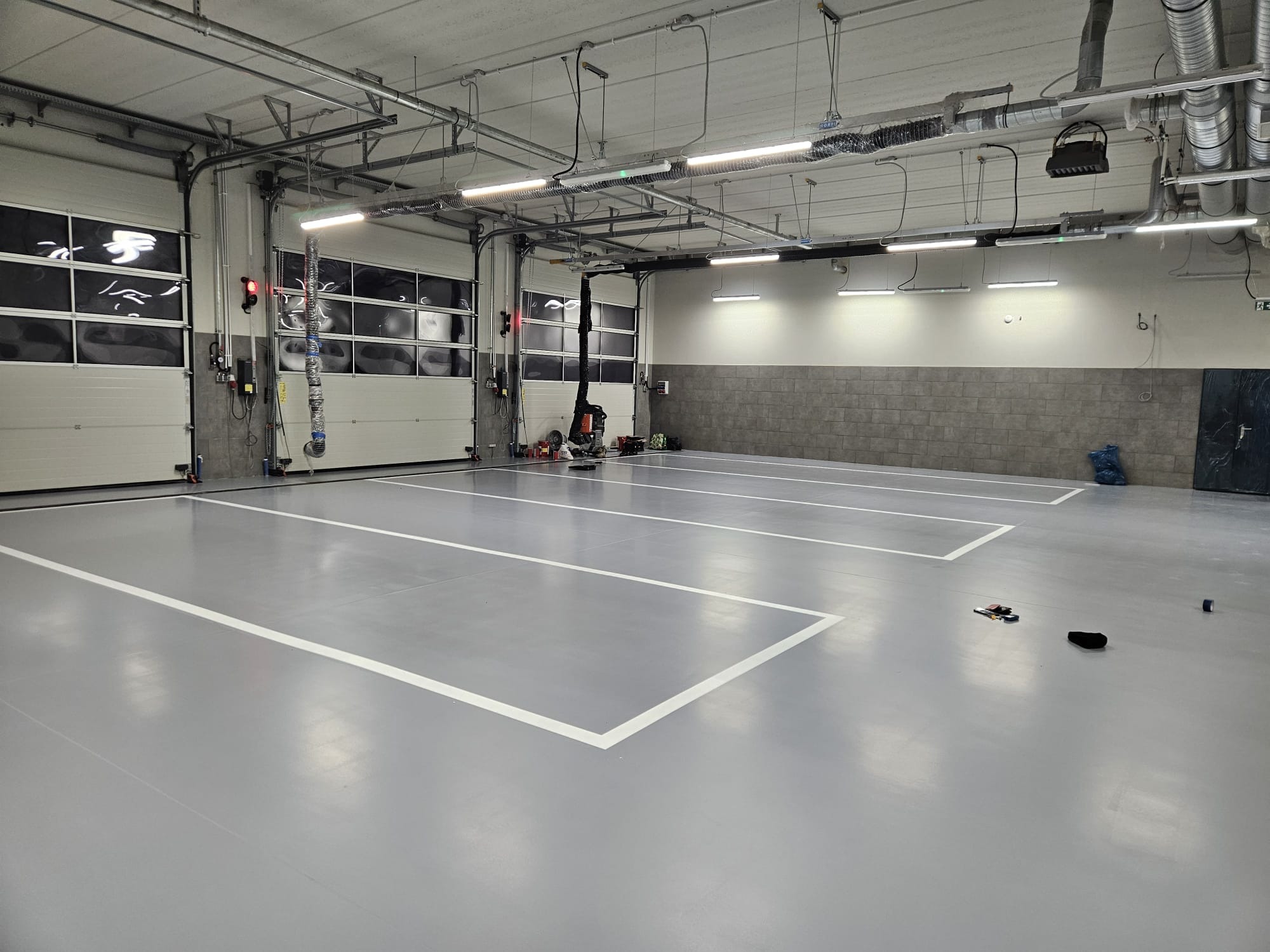 Szara, gładka podłoga w hali z wyznaczonymi białymi liniami stanowisk. Widoczne bramy garażowe, oświetlenie sufitowe i instalacje. Minimalistyczny, przemysłowy styl.