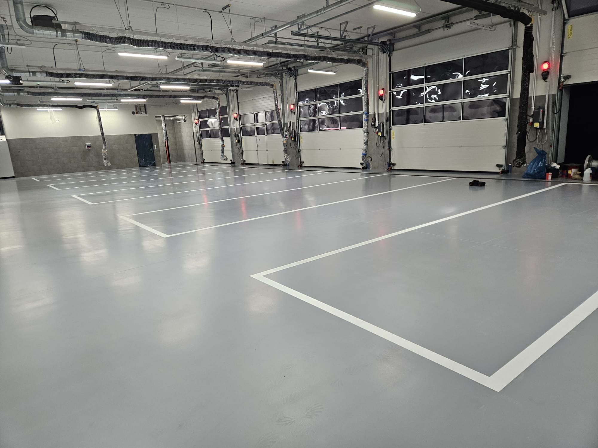 Jasnoszara posadzka przemysłowa z białymi liniami wyznaczającymi miejsca parkingowe w hali z bramami segmentowymi i instalacjami pod sufitem.