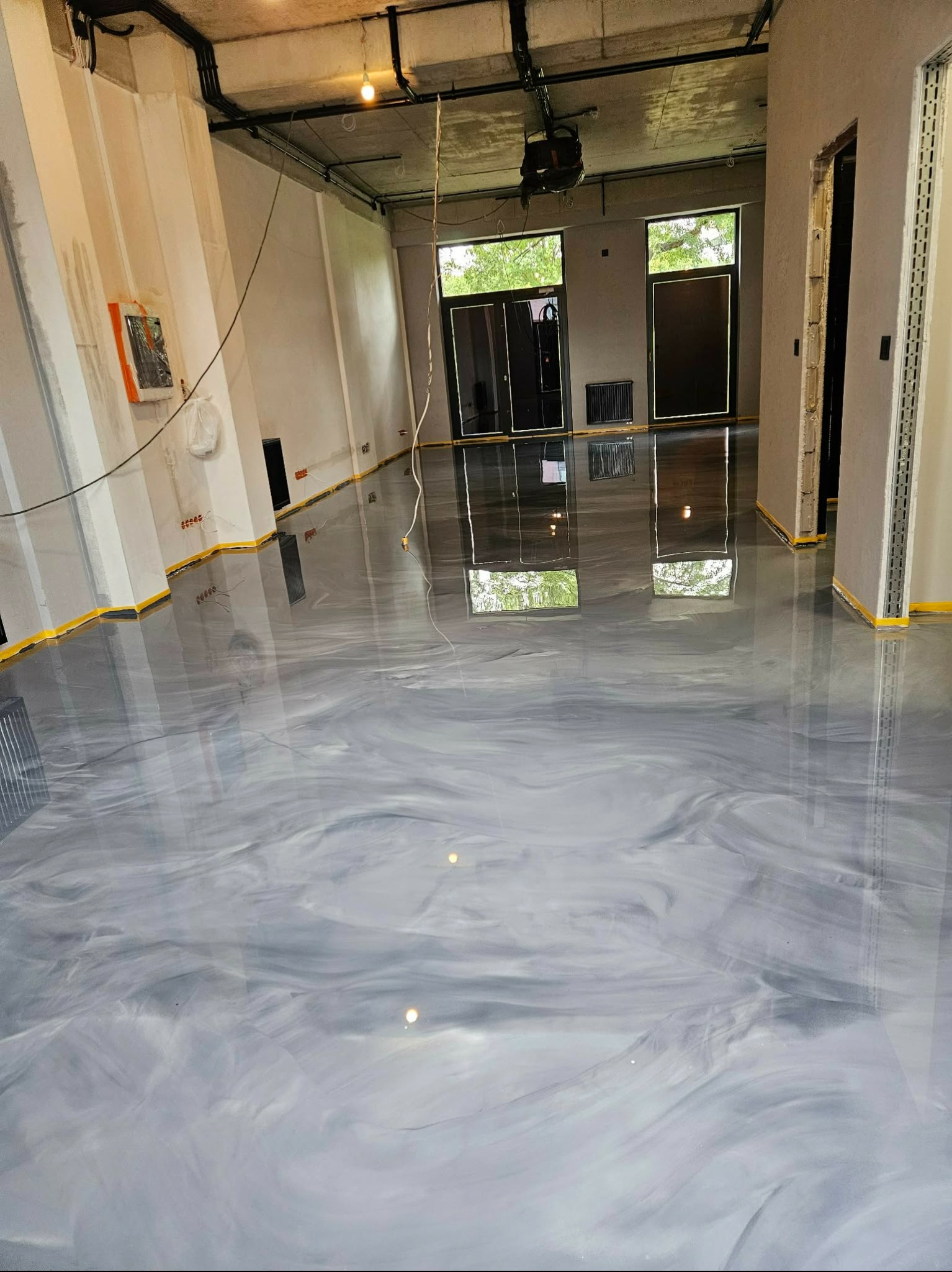 Błyszcząca, szara podłoga z efektem marmuru w przestronnym wnętrzu. Widoczne okna z odbiciem zieleni. Żółta taśma zabezpieczająca przy ścianach.