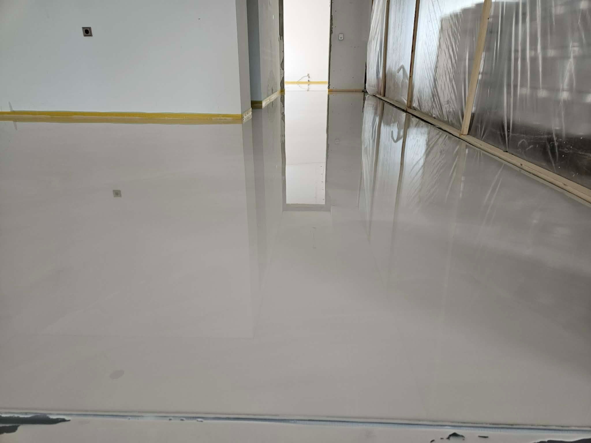 Błyszcząca, jasnoszara posadzka w nowym wnętrzu. Widoczne zabezpieczenia ścian folią i taśmą. Gładka powierzchnia odbija światło, tworząc efekt lustra.