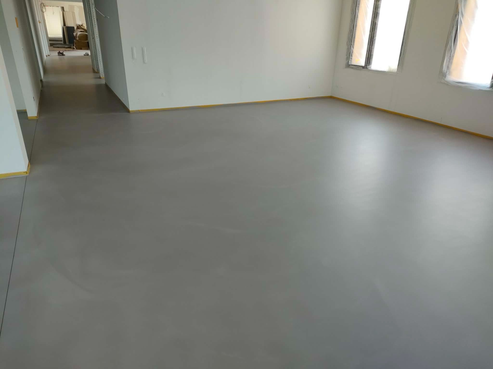Szara, gładka podłoga w jasnym pomieszczeniu z oknami zabezpieczonymi folią. Żółta taśma malarska wzdłuż ścian. Widok na dalsze pomieszczenia w głębi.