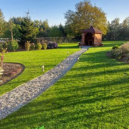 Robert Pilecki Consulting - Urokliwy ogród z zielonym trawnikiem, ścieżką z jasnego żwiru prowadzącą do drewnianej altany. W tle drzewa i krzewy, oświetlone popołudniowym słońcem.