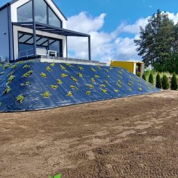 Robert Pilecki Consulting - Uformowany nasyp ziemny zabezpieczony czarną geowłókniną z nasadzonymi roślinami, na tle nowoczesnego domu z pergolą i błękitnego nieba z obłokami.