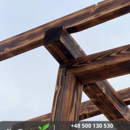 Robert Pilecki Consulting - Drewniana konstrukcja z belek o wyraźnym usłojeniu, złączona pod kątem, widziana z dołu na tle jasnego nieba. Element architektury ogrodowej w ciepłej tonacji brązu.