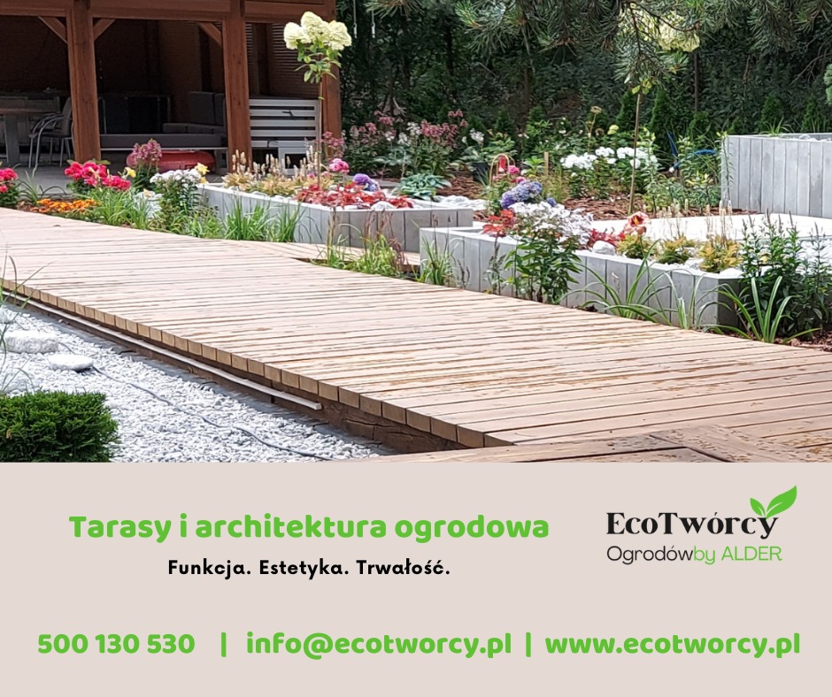 Drewniany taras prowadzący przez ogród z rabatami kwiatowymi w Janowcu Wielkopolskim. Betonowe obrzeża rabat, żwirowe podłoże, zieleń i kwiaty tworzą harmonijną całość.