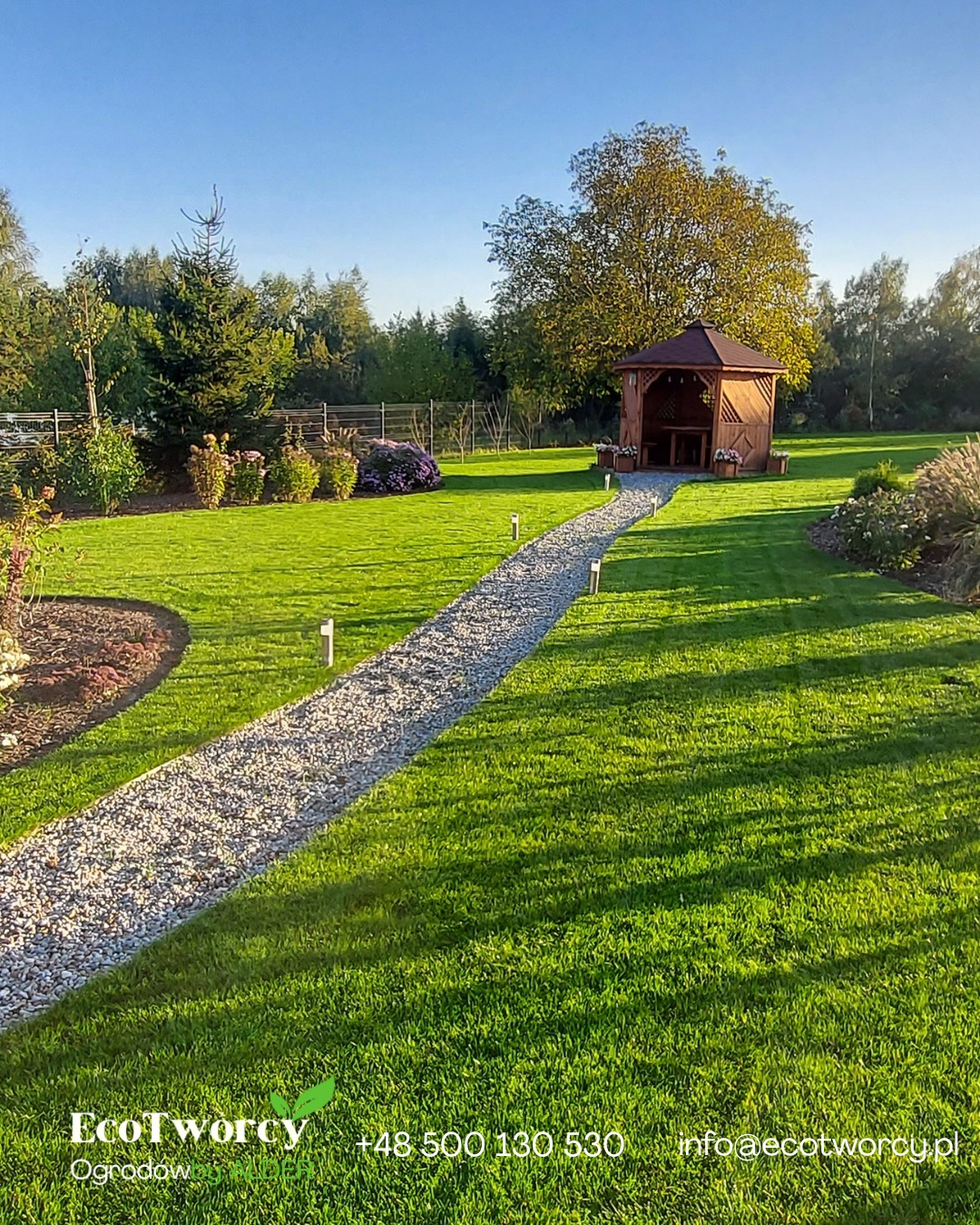 Urokliwy ogród z zielonym trawnikiem, ścieżką z jasnego żwiru prowadzącą do drewnianej altany. W tle drzewa i krzewy, oświetlone popołudniowym słońcem.