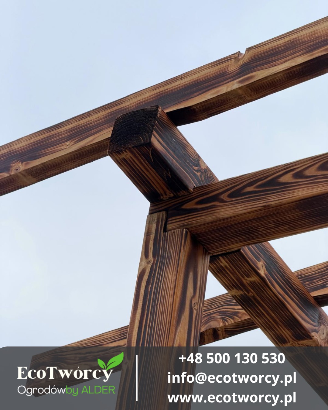Drewniana konstrukcja z belek o wyraźnym usłojeniu, złączona pod kątem, widziana z dołu na tle jasnego nieba. Element architektury ogrodowej w ciepłej tonacji brązu.