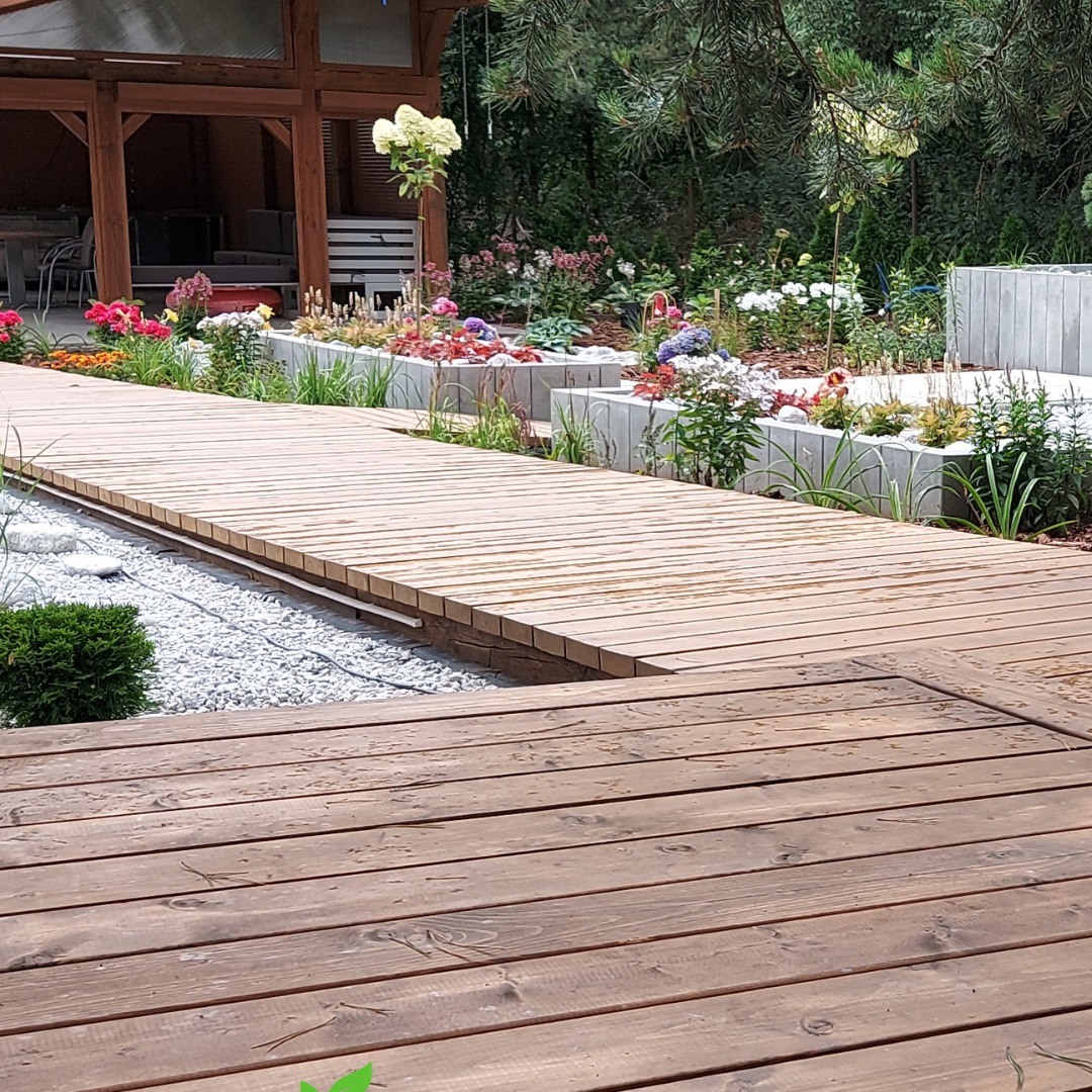 Drewniany pomost prowadzi przez ogród z rabatami kwiatowymi i białym żwirem, obok altana. Ujęcie z niskiej perspektywy podkreśla strukturę drewna.