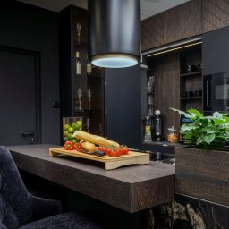 Duo s.c - Nowoczesna, czarna kuchnia z drewnianymi akcentami. Na blacie deska z pieczywem i warzywami, obok rośliny w donicy. Widoczny okap i zabudowa kuchenna.