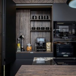 Duo s.c - Nowoczesna, ciemna kuchnia z drewnianymi akcentami. Widoczne zabudowane sprzęty, półki z naczyniami i ekspres do kawy. Blat z ciemnego kamienia. Oświetlenie LED.