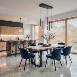 Duo s.c - Nowoczesna jadalnia z drewnianym stołem, aksamitnymi krzesłami i designerską lampą wiszącą. W tle widoczna kuchnia z wyspą i hokerami, całość w jasnych tonacjach.