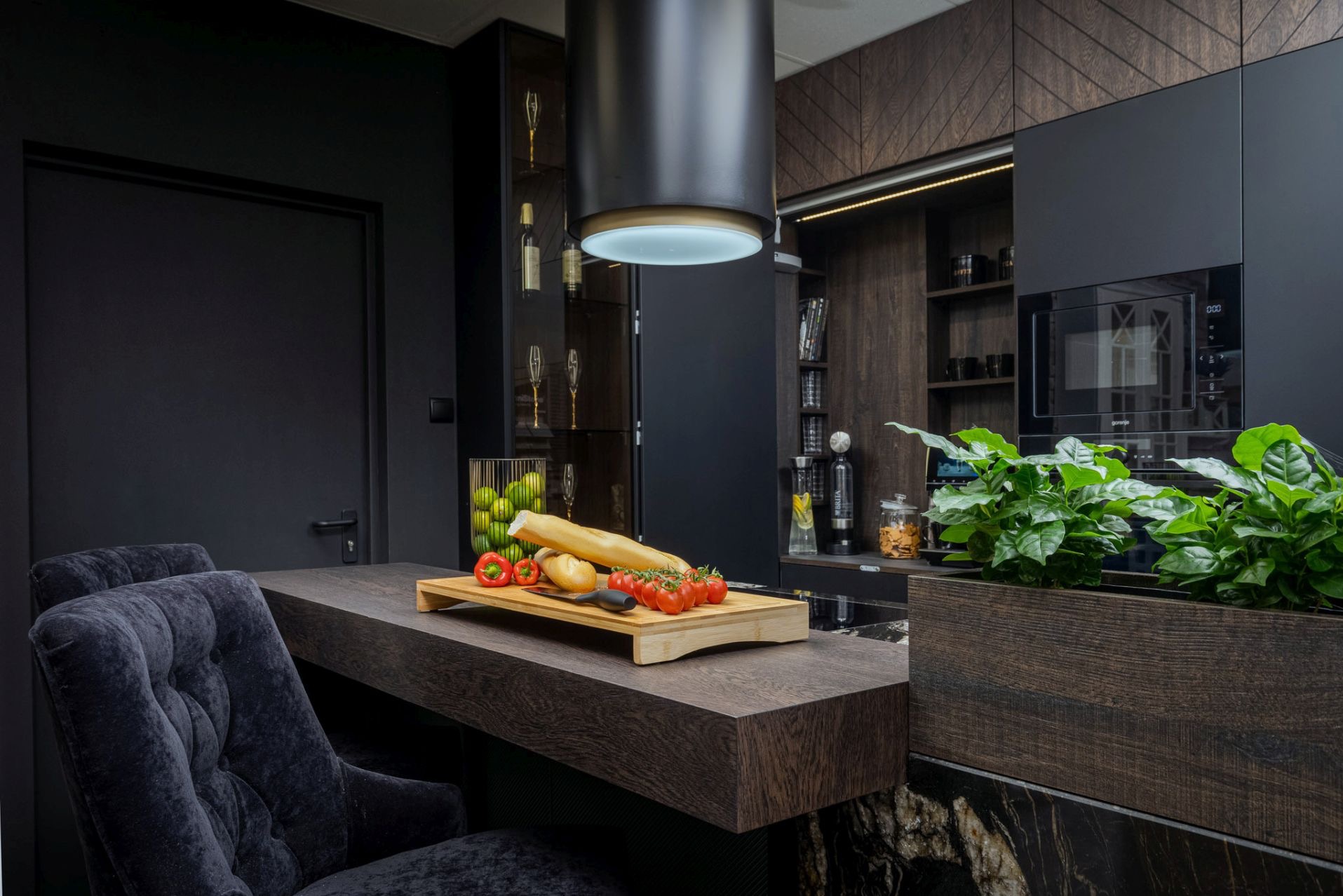 Nowoczesna, czarna kuchnia z drewnianymi akcentami. Na blacie deska z pieczywem i warzywami, obok rośliny w donicy. Widoczny okap i zabudowa kuchenna.
