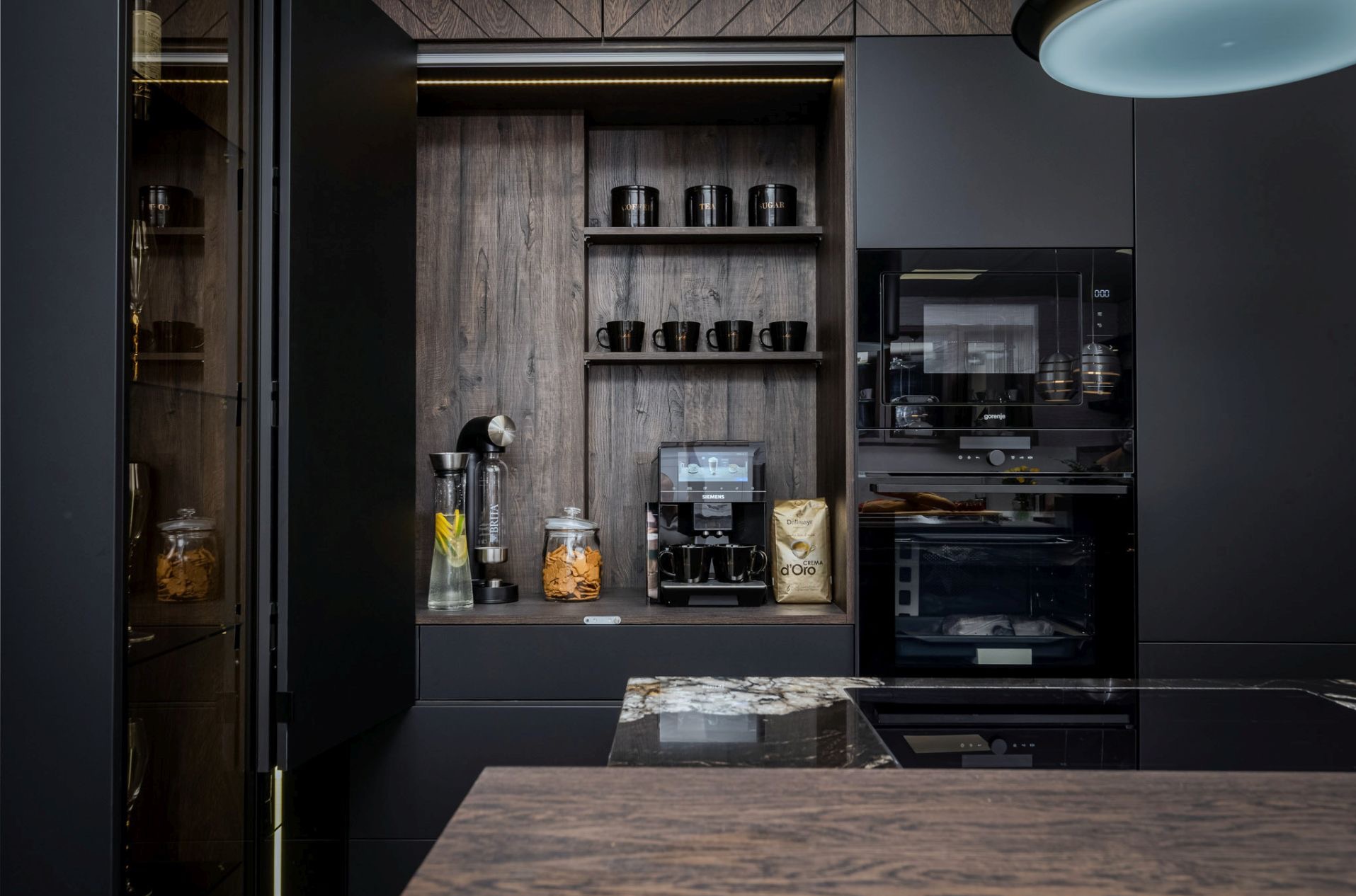 Nowoczesna, ciemna kuchnia z drewnianymi akcentami. Widoczne zabudowane sprzęty, półki z naczyniami i ekspres do kawy. Blat z ciemnego kamienia. Oświetlenie LED.