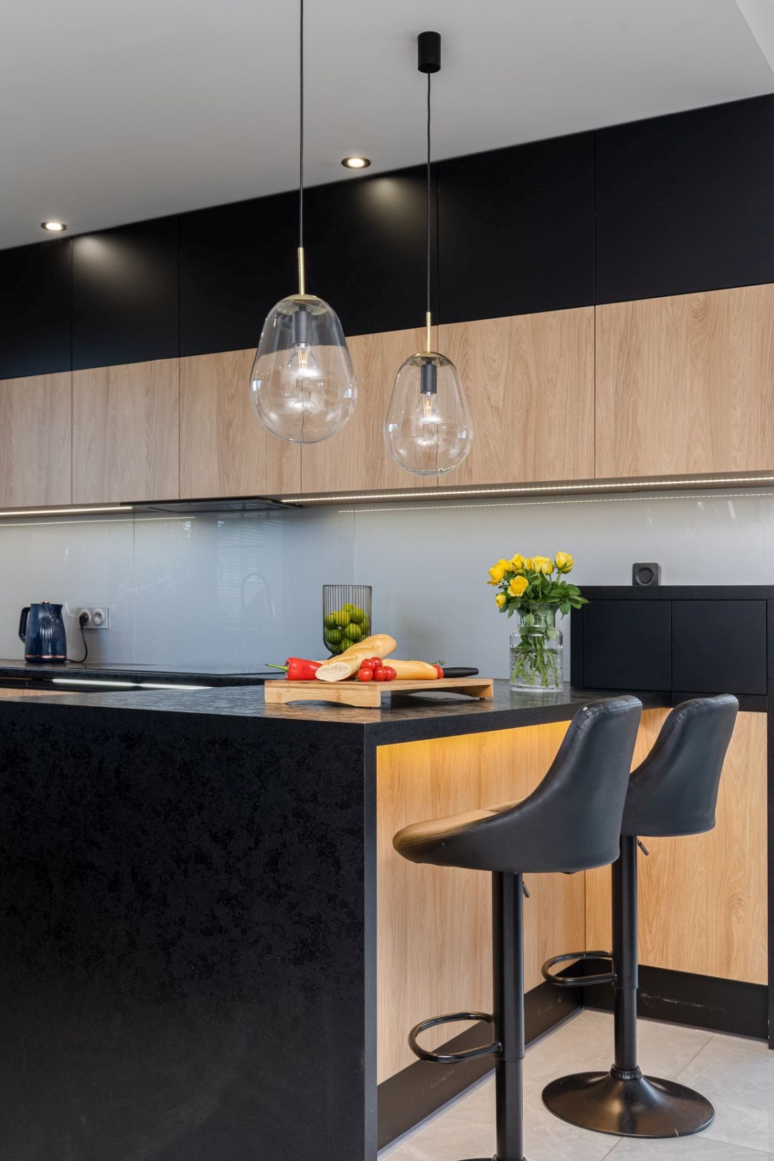 Nowoczesna kuchnia z wyspą: czarny blat, drewniane szafki, szklane lampy wiszące. Na blacie deska z pieczywem i warzywami, kwiaty w wazonie. Dwa czarne hokery.