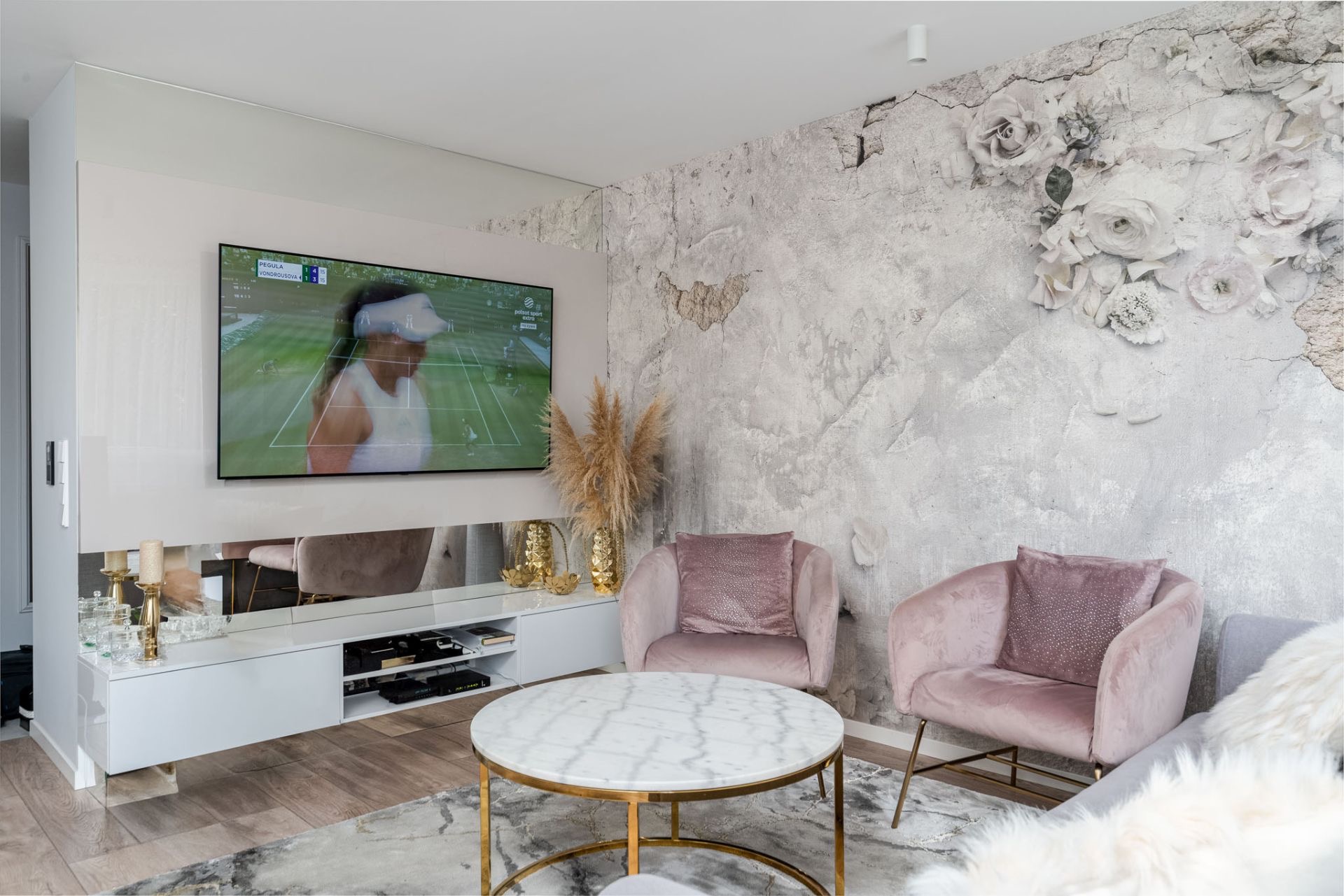 Salon z telewizorem, białą szafką RTV z lustrzanymi frontami, marmurowym stolikiem i fotelami w odcieniach różu. Na ścianie dekoracyjny tynk z motywem kwiatowym.