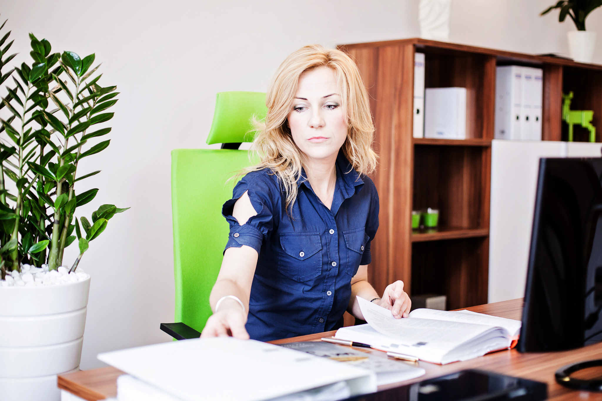 Kobieta w biurze, przegląda dokumenty na biurku z zielonym krzesłem. Na półce segregatory i dekoracje. Jasne wnętrze, skupiona twarz.