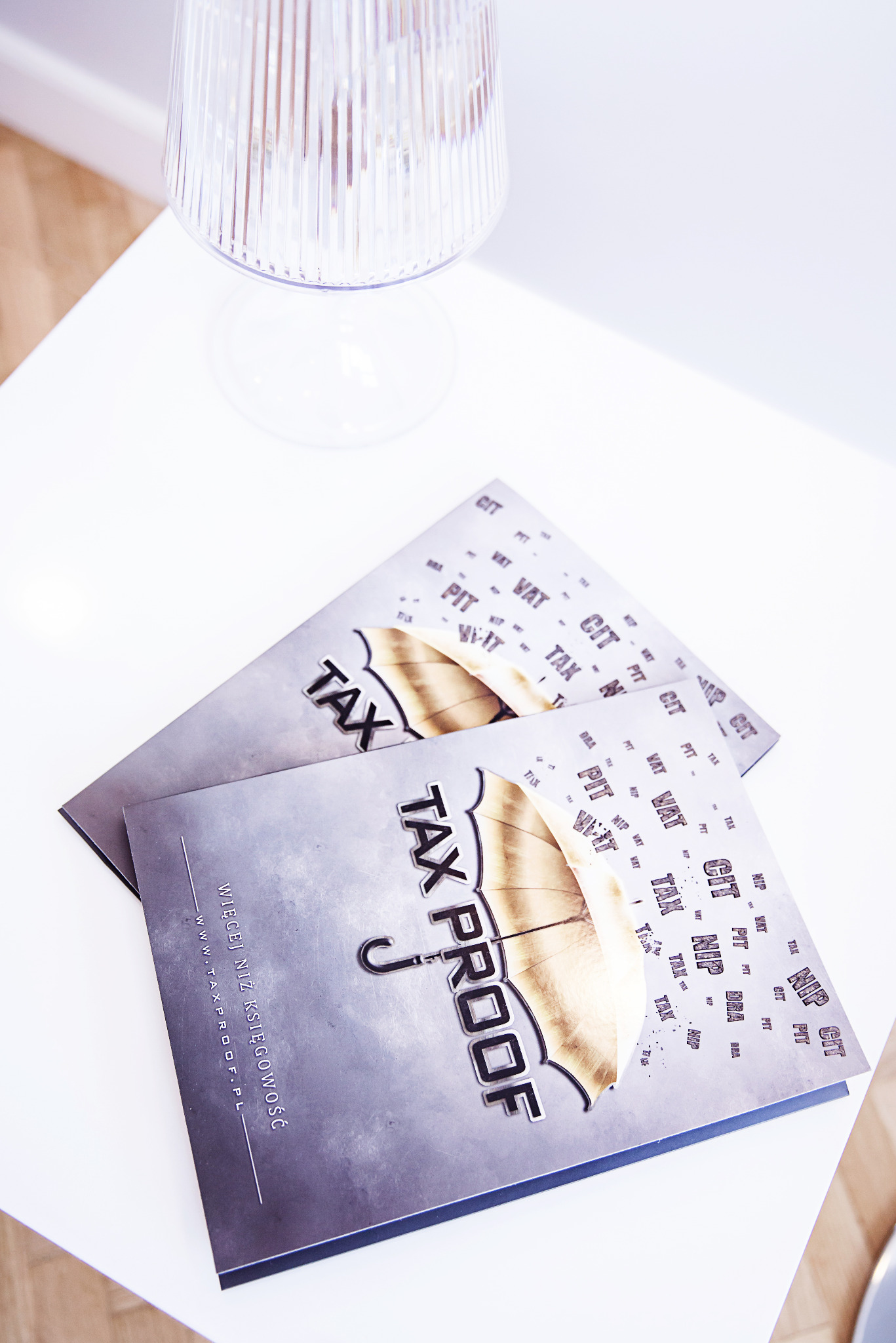 Dwa foldery informacyjne 'Tax Proof' leżące na białym stole, obok lampki. Foldery zawierają hasła związane z podatkami: VAT, CIT, PIT, NIP, TAX.