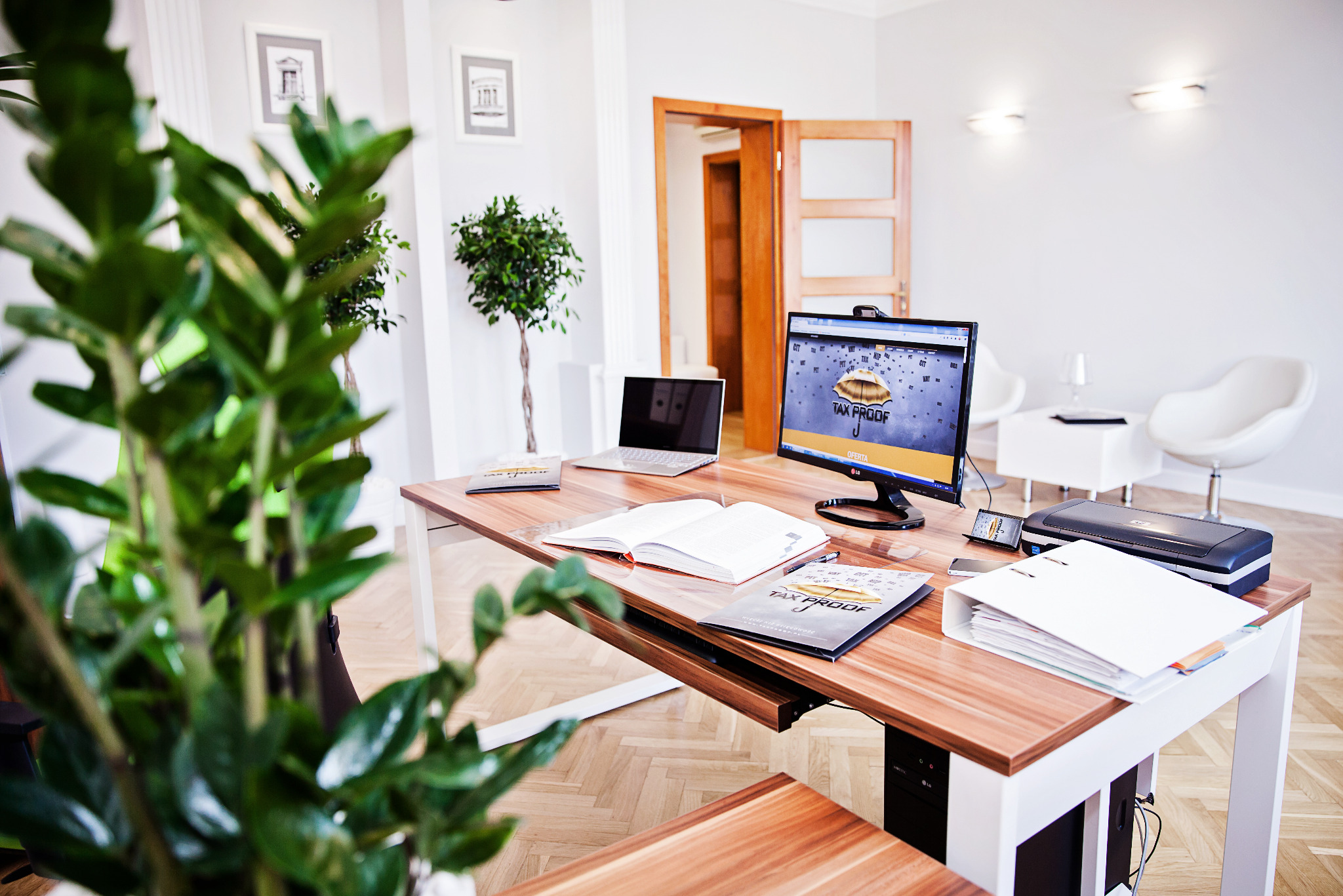 Nowoczesne biuro z drewnianym biurkiem, komputerem z logo firmy, roślinami i otwartą książką. Jasne wnętrze, minimalistyczny design.