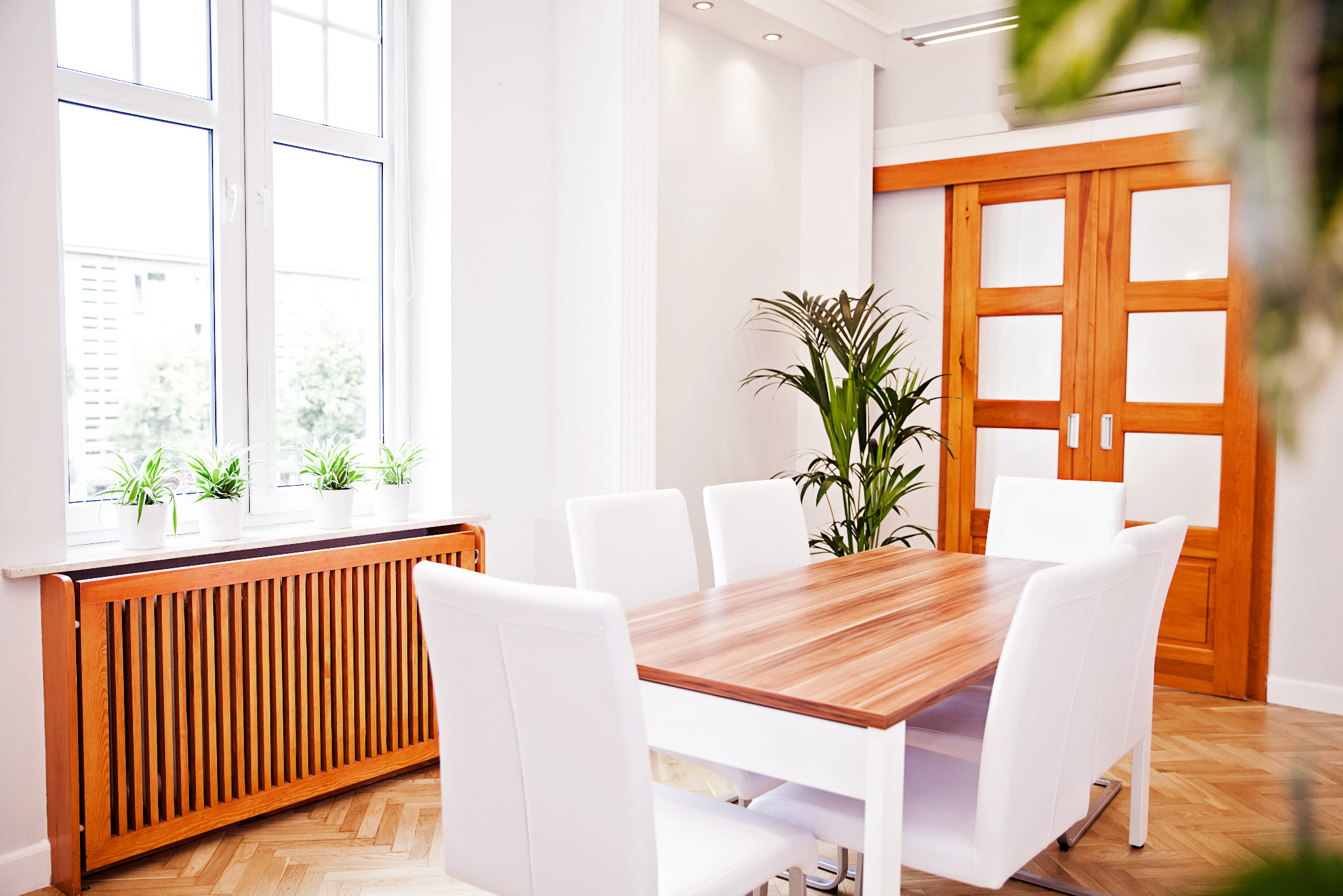 Jasne biuro z drewnianym stołem, białymi krzesłami i roślinami doniczkowymi. Widok na okno z zielenią na zewnątrz. Drewniane drzwi przesuwne w tle.