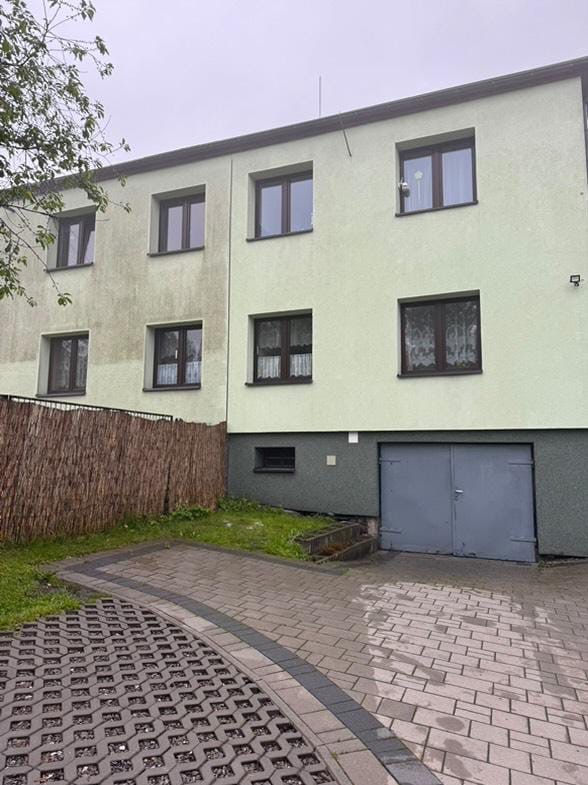 Dwukondygnacyjny budynek z elewacją w dwóch odcieniach zieleni, z widoczną różnicą w czystości. Kostka brukowa przed budynkiem. Szara brama garażowa.