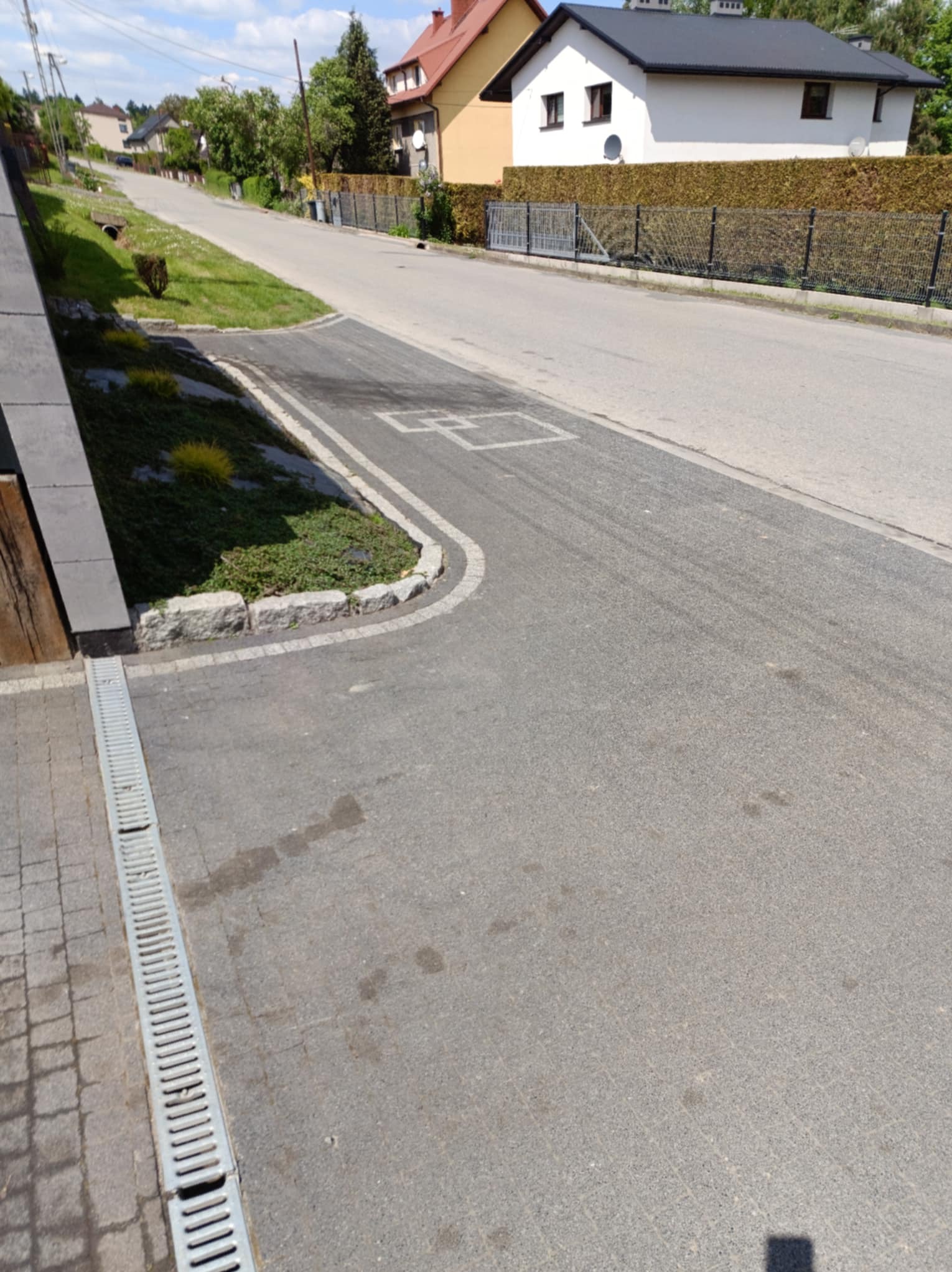 Ulica z kostki brukowej z odpływem liniowym, obrzeżem z kamienia i zielenią. W tle domy jednorodzinne i błękitne niebo.