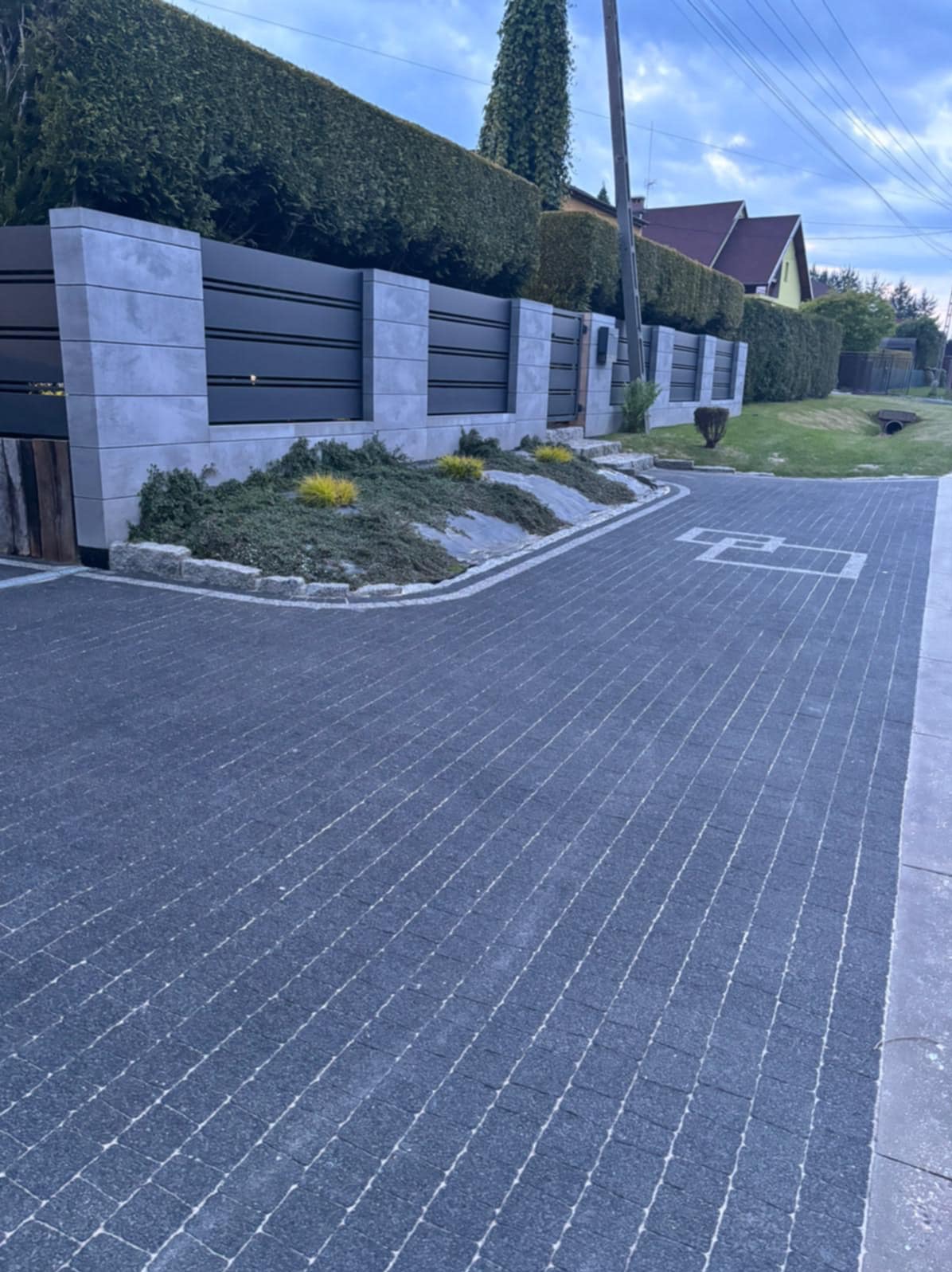 Ułożona, ciemnoszara kostka brukowa z jasnymi fugami, tworząca wzór. W tle nowoczesne ogrodzenie z elementami betonowymi i zielenią.