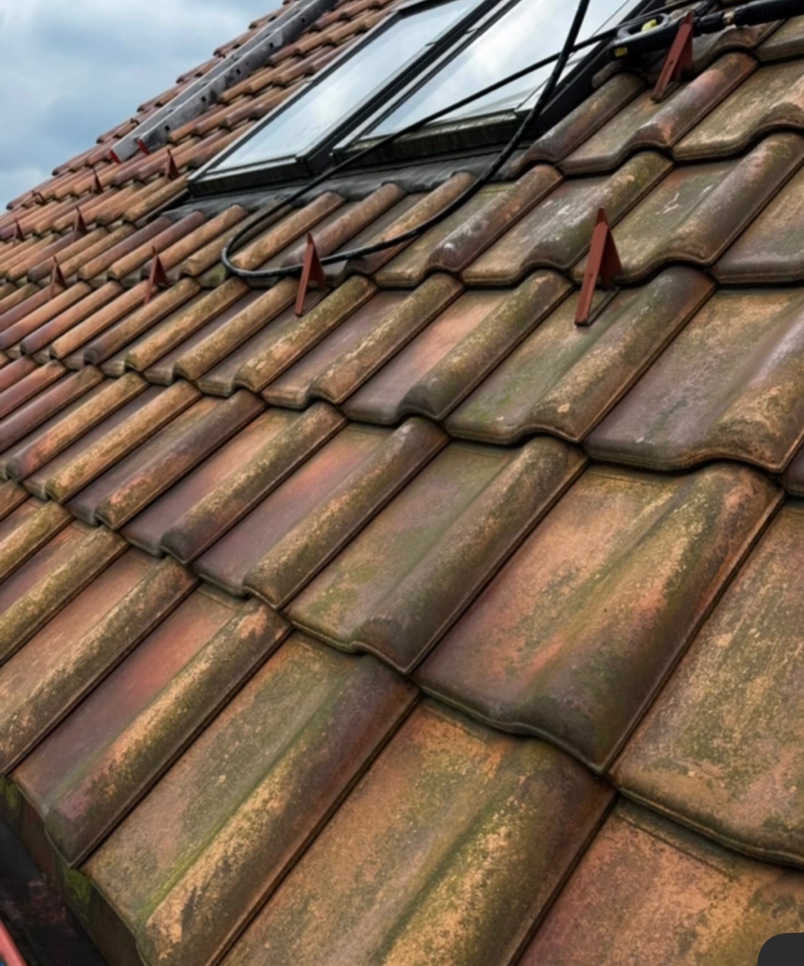 Czerwono-brązowa dachówka pokryta mchem, z zamontowanymi oknami dachowymi i zabezpieczeniami. Widoczna drabina i przewody na dachu.