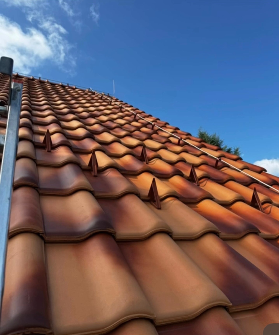 Czerwono-brązowy dach z dachówki ceramicznej, widziany pod kątem z dołu, z fragmentem drabiny po lewej stronie i błękitnym niebem w tle.