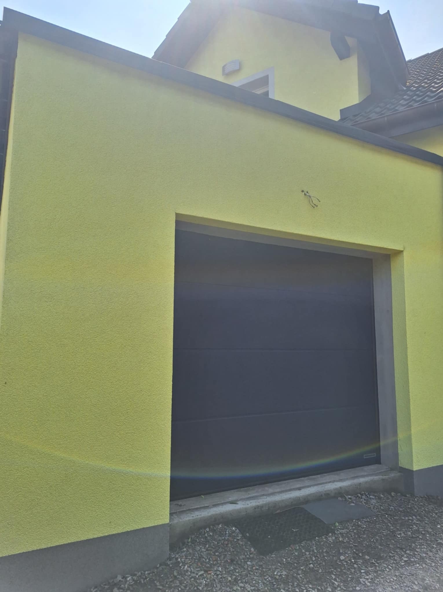 Żółta elewacja budynku z szarą bramą garażową. Widoczne ślady po instalacji na ścianie. Ujęcie z dołu, w tle fragment dachu i niebieskie niebo.