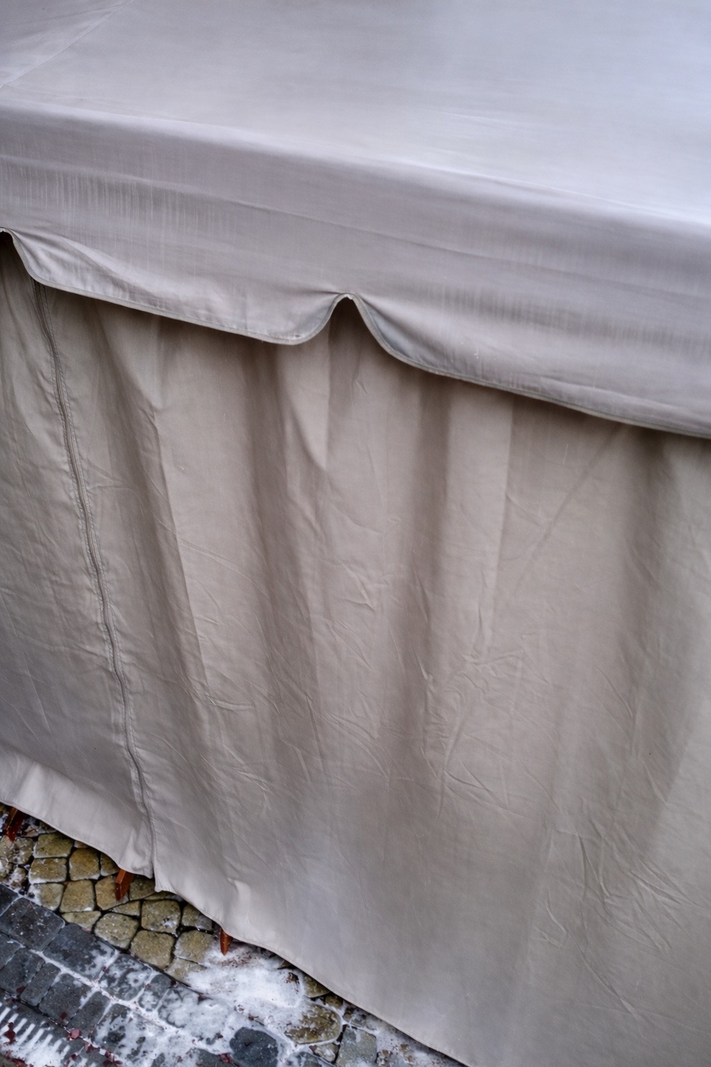 Beżowy, materiałowy baldachim z dekoracyjnym wykończeniem, częściowo zasłaniający kostkę brukową z resztkami śniegu. Widoczny fragment konstrukcji.