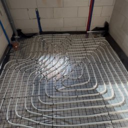 TKI Hydraulika Cezary Figiel - Białe rury ogrzewania podłogowego ułożone na czarnej siatce w pomieszczeniu z oknem dachowym i ścianami z białych bloczków. Widoczne niebieskie i czerwone rury.