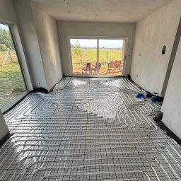 TKI Hydraulika Cezary Figiel - Białe rury ogrzewania podłogowego ułożone na czarnej siatce w surowym wnętrzu z dużymi oknami i widokiem na zewnątrz. Widoczne narzędzia i materiały instalacyjne.