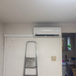 TKI Hydraulika Cezary Figiel - Biały klimatyzator Kaisai zamontowany na ścianie, widoczna drabina i fragment kuchni w tle. Na wyświetlaczu klimatyzatora widoczna temperatura 19 stopni.