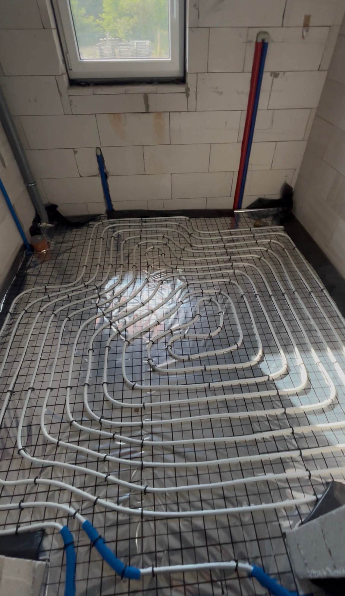 Białe rury ogrzewania podłogowego ułożone na czarnej siatce w pomieszczeniu z oknem dachowym i ścianami z białych bloczków. Widoczne niebieskie i czerwone rury.