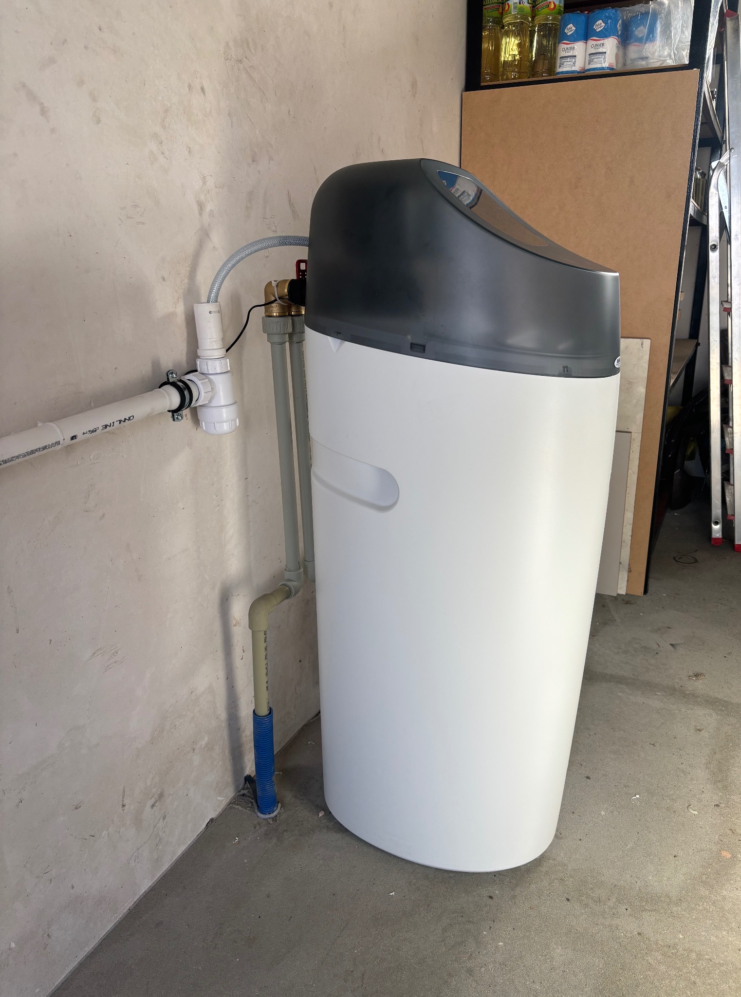 Biały zmiękczacz wody z szarą górną częścią, podłączony do instalacji hydraulicznej na tle surowej ściany i regału z zapasami.