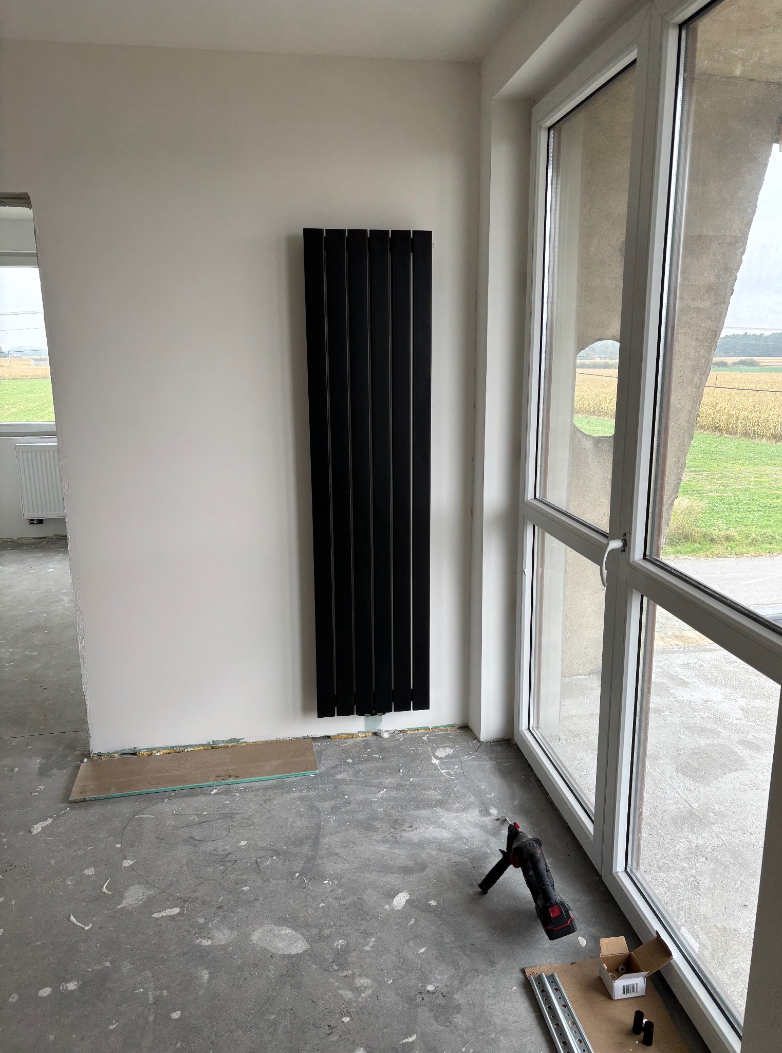Czarny, pionowy grzejnik panelowy zamontowany na białej ścianie w Gostyninie. Obok okno z widokiem na pole. Na podłodze narzędzia i akcesoria montażowe.
