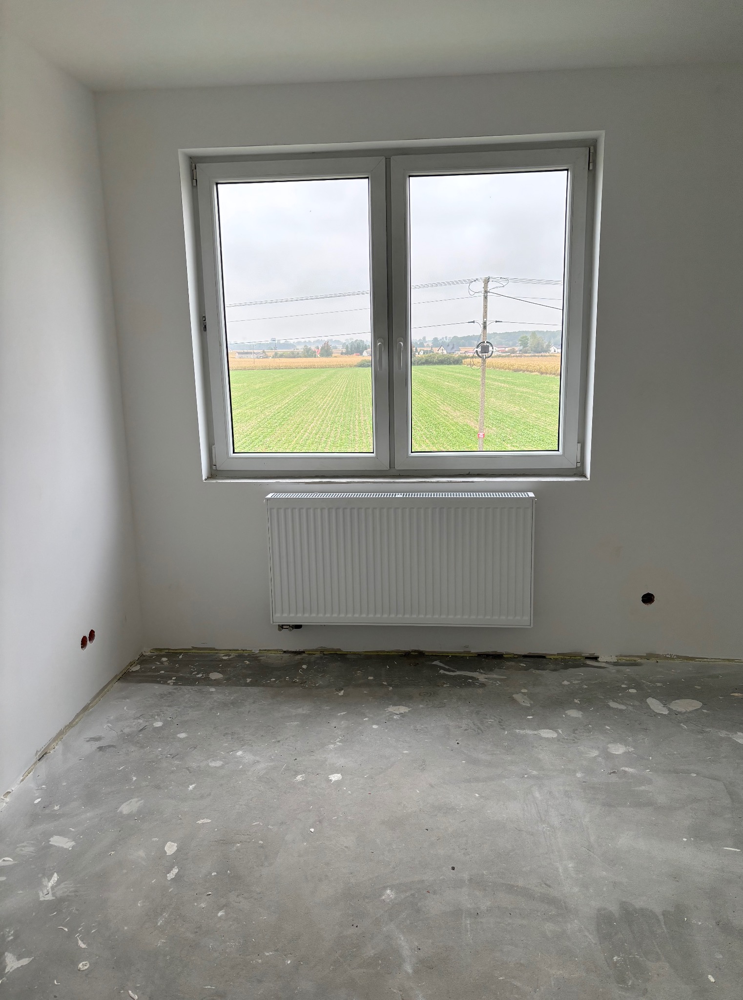 Biały grzejnik panelowy pod oknem w surowym, jasnym pomieszczeniu z widokiem na pole. Przygotowane punkty instalacji na ścianie.