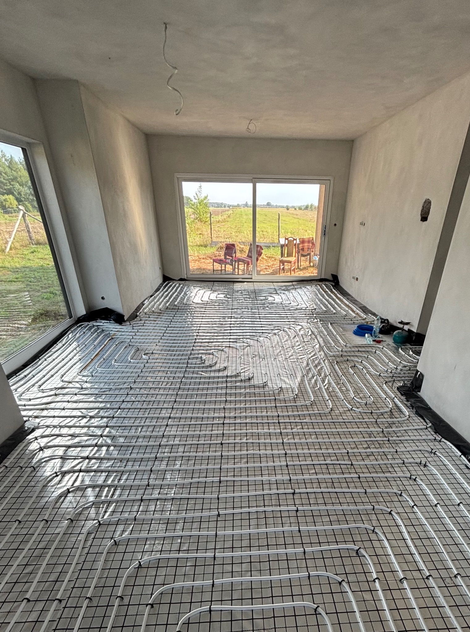 Białe rury ogrzewania podłogowego ułożone na czarnej siatce w surowym wnętrzu z dużymi oknami i widokiem na zewnątrz. Widoczne narzędzia i materiały instalacyjne.
