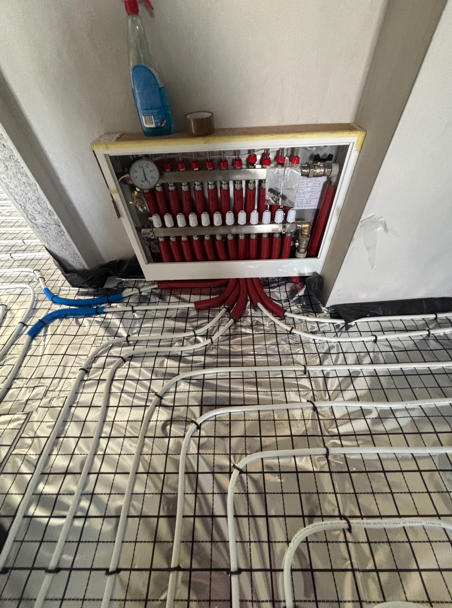 Rozdzielacz ogrzewania podłogowego z czerwonymi rurami, białe rury ułożone na srebrnej folii z czarną siatką, widok z góry, instalacja w trakcie montażu.