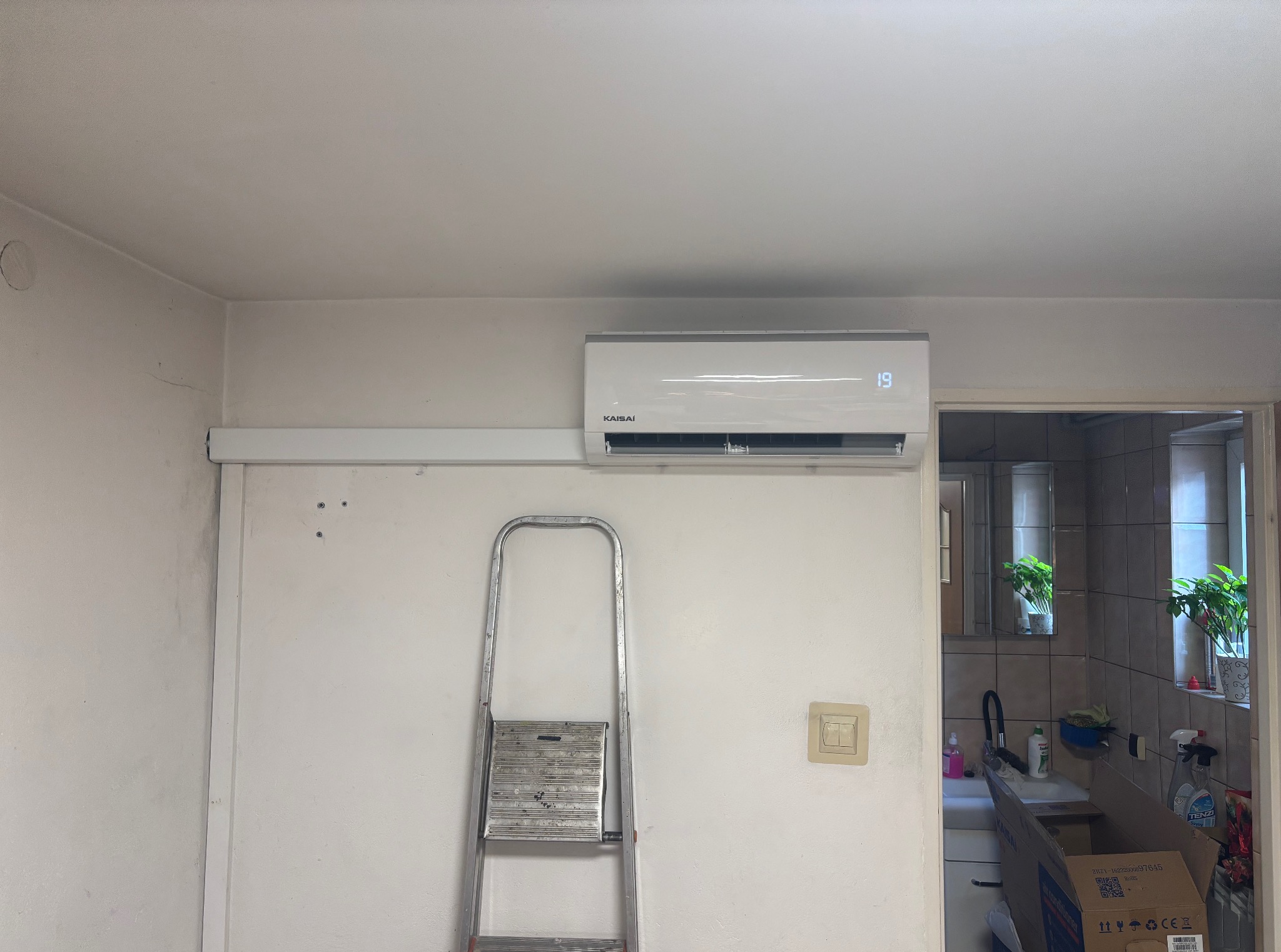 Biały klimatyzator Kaisai zamontowany na ścianie, widoczna drabina i fragment kuchni w tle. Na wyświetlaczu klimatyzatora widoczna temperatura 19 stopni.