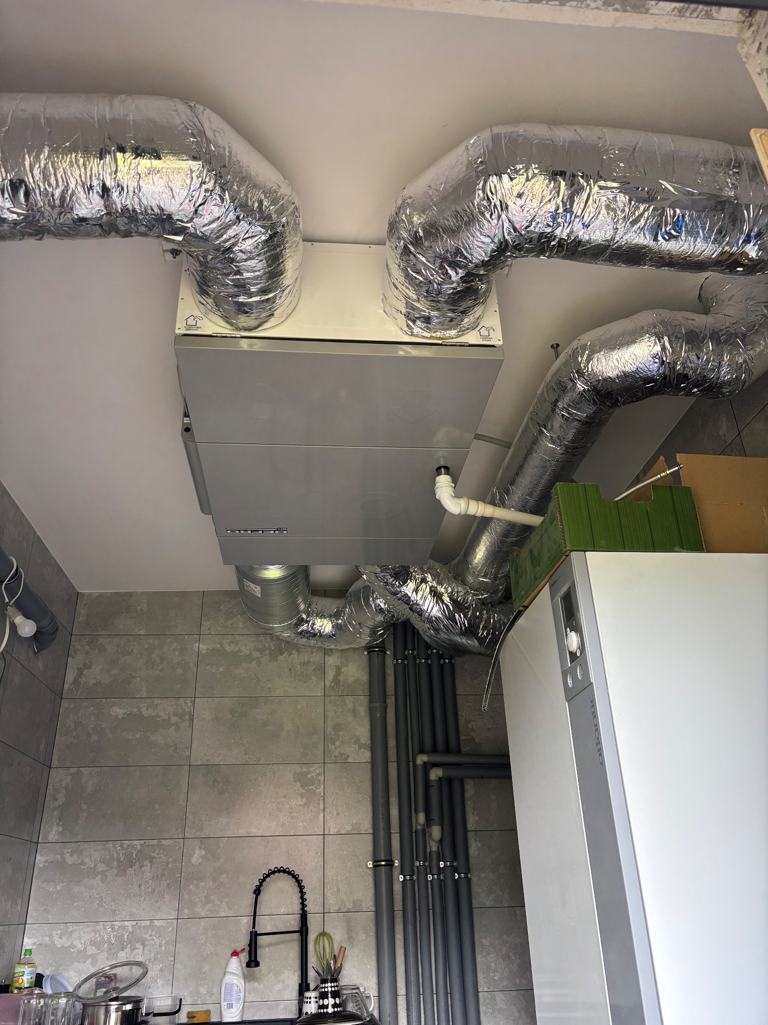 Szare urządzenie rekuperacji pod sufitem, z podłączonymi, izolowanymi przewodami wentylacyjnymi. Widoczne rury i fragment kotła grzewczego w pomieszczeniu z szarą glazurą.