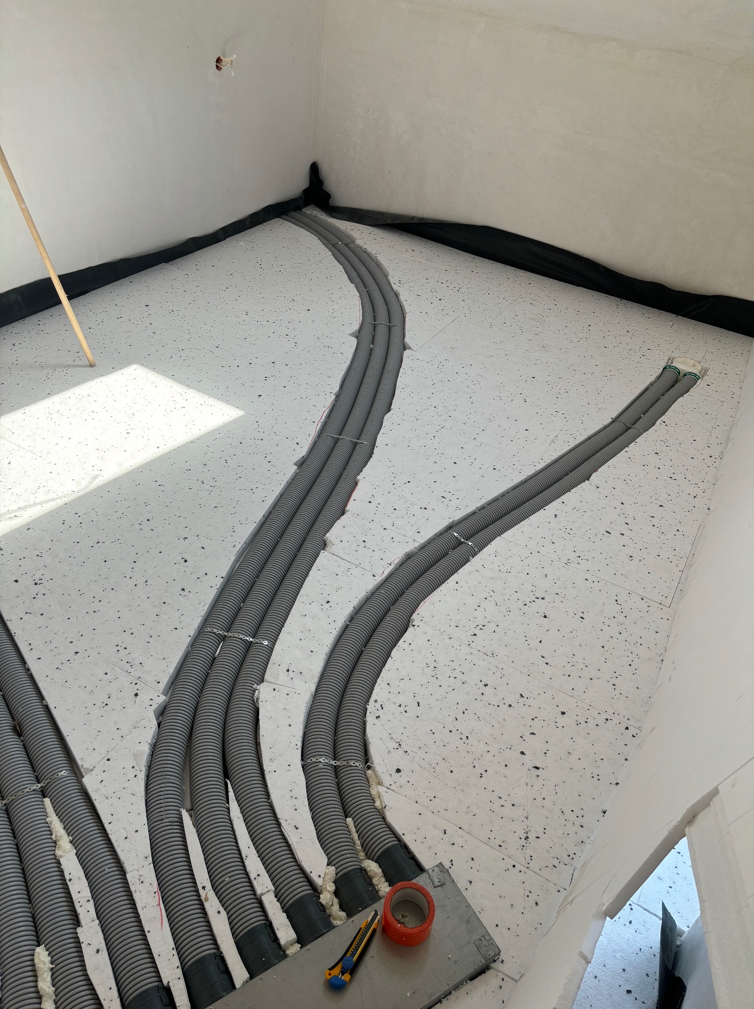 Układanie szarych rur w wycięciach styropianu pod ogrzewanie podłogowe. Widoczne narzędzia i taśma. Czarna folia przy ścianach. Jasne wnętrze w trakcie prac instalacyjnych.